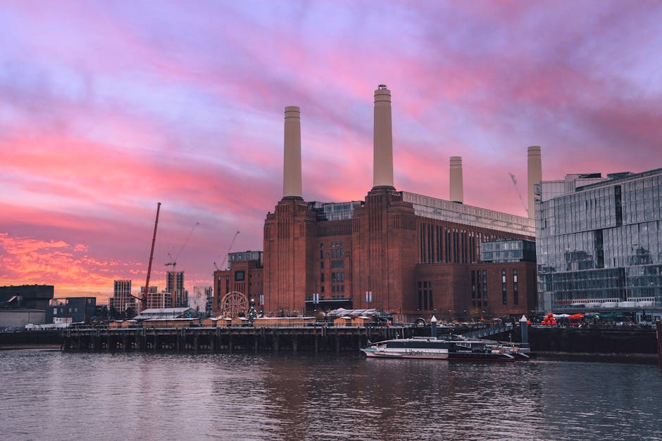 A scenic view of the Battersea Power Station with its iconic four tall chimneys, set against a vibrant pink and purple sunset sky. In the foreground, the River Thames reflects the colorful sky and the