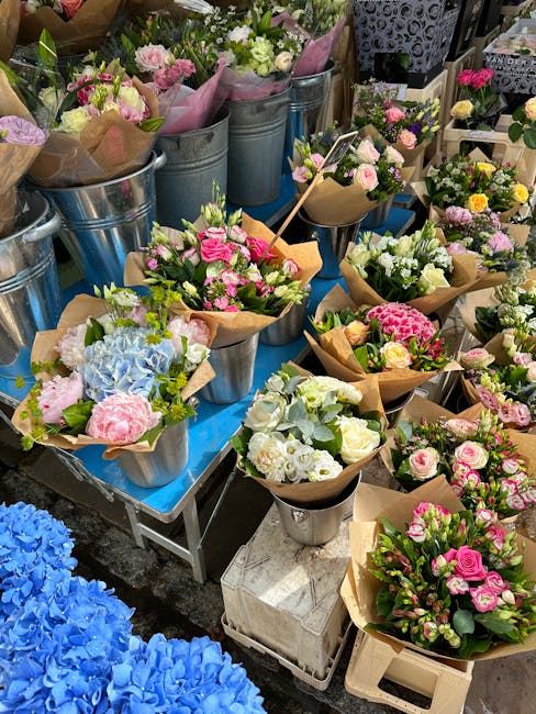 A vibrant display of floral arrangements and bouquets on a table at a flower market. The flowers include pastel pink roses, white lisianthus, pale yellow blooms, and soft blue hydrangeas, all arranged