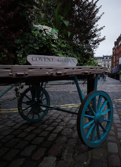 A vintage wooden cart with a blue wheel and frame, positioned on a cobblestone street, filled with a lush arrangement of white and green flowers including hydrangeas, roses, and foliage, set against a