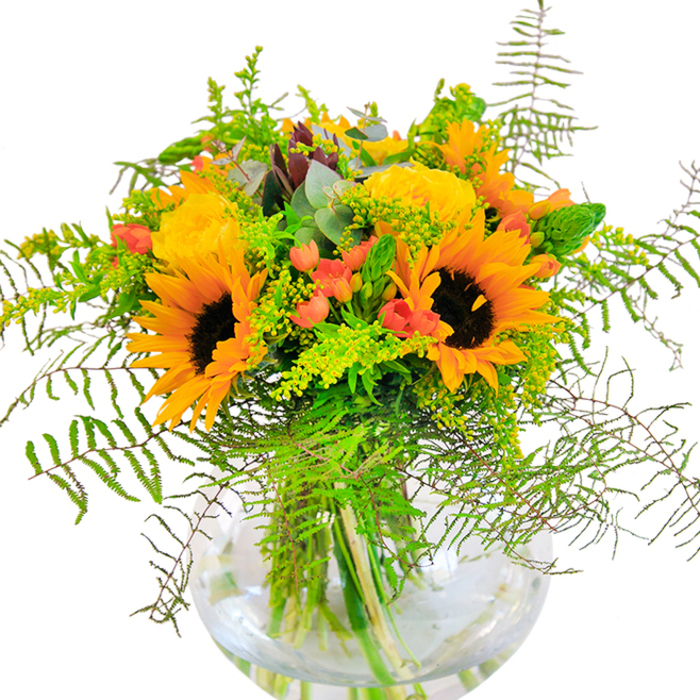 From the first glance, this luminous arrangement conjures the spirit of Nine Elms in late summer, bursting with radiant hues of yellow and orange that echo the vibrant community and sunlit Thames-side strolls. Sunflowers unfurl their wide, golden petals, each centered by a deep brown disk, exuding a confident optimism that brightens any occasion. Nestled among them, garden-fresh yellow roses reveal layers of velvety, spiraled petals-classic symbols of appreciation and joy, reminiscent of the blooms found at the nearby New Covent Garden Flower Market. Crimson Hypericum berries dot the bouquet, their glossy surfaces providing dynamic contrast against a background of lush, textured foliage. Feathery fern fronds drape gracefully, while sprigs of goldenrod and the muted blue-green tones of eucalyptus introduce a contemporary, breezy element, reflecting the mixture of modernity and greenery that defines Nine Elms' evolving landscape. All these elements are artfully arranged within a clear, bubble-shaped glass vase, the stems left exposed, cascading in natural lines and illuminated by the pure, clean light of a new day. The arrangement exudes a subtle, uplifting sweetness-its blend of lively colour, gentle movement, and harmonious composition making it ideal for transforming any living space, Thames-side office, or rooftop garden gathering. Crafted by local Nine Elms Florist artisans, this bouquet is perfect for celebrating milestones, sharing happiness, or adding a heartfelt touch to spaces that matter most throughout the neighbourhood.